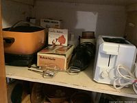 Photo showing orange crock pot, boxed Robot Marie immersion blender, hand mixer beaters, white two-slice toaster, and boxed Black & Decker chopper on a shelf