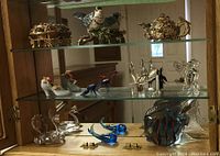 Photo showing shelves with multiple items including gold-tone trinket boxes, ceramic bird shoes, glass bird figurines, crystal peacock, glass fish, and clear glass swan figurines.