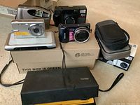 Lot of 6 cameras and 2 camera cases arranged on boxes and floor, showing different brands and models.