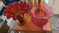 Photo of two cranberry glass vases on wooden surface, one ruffle style, one rounded with ruffled top, providing overall view.