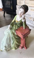 View of Royal Doulton Carolyn porcelain figurine from the front and right side, showing seated woman in green dress with reddish-brown side table.