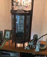 Wooden curio display cabinet with glass panels and several figurines arranged outside and inside, including dog and bird figurines, with a glass bell on top and framed artwork nearby.