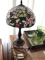 Front angled view of the lamp showing the marbled resin stained glass style floral shade and bronze base on a wooden surface with green glassware nearby.