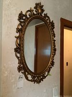 Ornate bronze-toned oval wall mirror mounted on a wall with decorative molded frame showing floral and scroll details, photographed from the front and angles to show the frame design and condition.