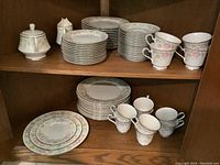 Photo showing stacked plates including three sizes and cream and sugar set alongside cups and saucers on wooden shelf.
