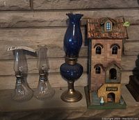 Three oil lamps and one wooden birdhouse on stone background, showing clear glass lamps, blue glass lamp, and cottage birdhouse