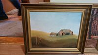 Framed oil painting showing a rural barn and landscape under blue sky placed on wooden floor.