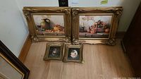 Four framed prints arranged on the floor, two large gold ornate frames with still life scenes and two smaller gold frames with floral images.