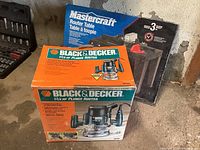 Two boxes side by side on concrete floor: Black & Decker plunge router box in front and Mastercraft router table box behind.