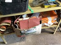 Box filled with various sandpaper sheets and sanding belts on lower garage shelf with other tools visible