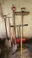 Photo showing five outdoor tools including two dirt shovels, tamper, garden rake, and red snow shovel standing against a wall.