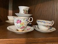 Photo of five teacup and saucer sets on wooden shelf showing variety of floral and scenic patterns.