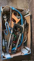 Top-down view of box containing various hand tools including saws, scissors, and utility blades arranged in a cardboard box.