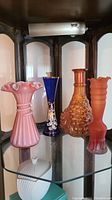 Four art glass vases displayed on glass shelf: pink ruffled top, cobalt blue floral, amber bubble pattern, and orange frosted with ribbing.