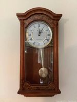 Front view of the Bulova wall clock showing the entire wooden frame, clock face with Roman numerals, and pendulum behind glass.