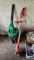 Weed trimmer standing upright beside the leaf blower placed on a black pail, showing the full items in the lot.