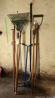 Lot of 5 yard tools including 2 rakes, manual tiller, hoe, and pitchfork leaning against a wall on garage floor.