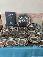 Wide view of 20 pewter Christmas plates with embossed designs by Astri Holthe laid out on a table with a matching pewter clock behind them.