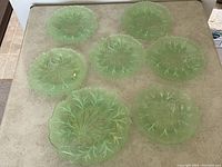 Seven green depression glass plates arranged on a table, showcasing the textured leaf design pattern on each plate, one larger plate and six smaller plates.