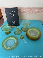Full set of green glass items including luncheon plates, taper candle holders, and candy bowls displayed on a blue surface, along with a black folder marked 'Lot 52'.