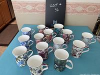 Overall view of 13 floral design Maxwell cups in various colors and flower patterns on a blue tablecloth.