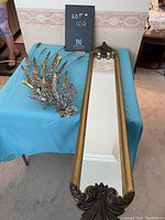 Photo showing the long gold-coloured framed mirror laying flat on a blue tablecloth alongside decorative gold-coloured metal wall decor with feather pattern.