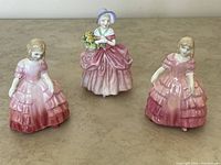Front view of three Royal Doulton figurines: two versions of "Rose" in pink gowns and one "Cissie" with bonnet and basket of flowers