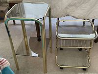 Two brass finish occasional tables with glass tops, one rectangular with glass shelf, the other square with chrome metal rack on wheels, shown on carpet and under bed.