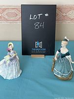Photo of two Royal Doulton figurines named Clare and Clarenda on blue cloth with a black sign behind them marked Lot 84.