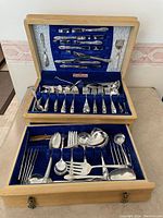 Full view of the open wooden cutlery case showing two tiers with multiple silver plate pieces organized in blue velvet compartments.