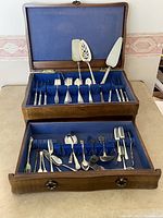 Full view of the wooden case with cutlery in two layers lined in blue fabric. The top section holds larger serving utensils, lower drawer holds an assortment of forks, spoons, knives and specialty cutlery.