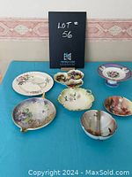 Top view of seven assorted decorative and serving dishes arranged on a teal cloth with a LOT 56 sign behind, showing varied shapes, sizes, and floral designs.