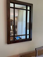 Brown wood framed wall mirror with mullions mounted on a light-colored wall, reflecting a chandelier, curtains, and furniture in a room.
