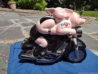 Side view showing two pink pigs, one with a visible tattoo, riding a black motorcycle on a stone patio with blue cloth underneath.
