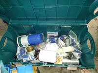 Green wheeled tote open, displaying assorted camping equipment including a blue and white thermos, metal containers, and small gear