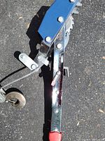 Photo of the main body of the come along hand winch showing the polished steel frame, ratchet gear, and caster wheel on asphalt.