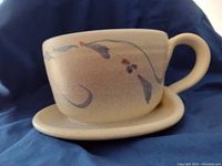 Soup bowl with attached handle and under plate on blue fabric background, side angle showing painted floral brushstroke design