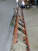 Two extension ladders leaning against a wall, one Werner orange fiberglass with aluminum rungs and the other an aluminum ladder with a wall attachment