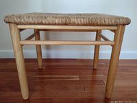 Side view of wicker seat showing woven wicker top and rounded wooden legs on hardwood floor.