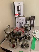 Photo showing coffee accessories set including boxed Vevok Chef manual coffee grinder, Secura milk frother, French press, Turkish coffee pot, cups, and saucers on a table.
