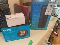 Photo showing Amazon Echo Spot box, Amazon Echo box and small white box with connecting cord on top, all on a white surface.