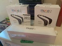 Two Muse 2 boxes in original plastic wrap behind one long white Muse S box with green tape reading “USB & Lightning cables”