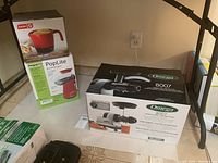 Three boxed appliances displayed on floor: PopLite hot air popper, Dash Express Hot Pot, Omega 8007 juicer