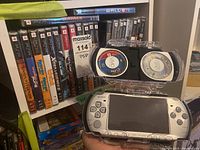 Row of sealed PSP UMD game and movie cases on shelf with open UMD case and PSP handheld in hand