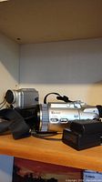 Wide shot of DCR-PC101 Handycam and Cyber-shot DSC-F717 cameras on shelf with battery charger