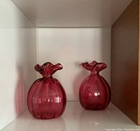 Two cranberry glass vases side by side inside a shelf, showing full front view and relative size.