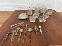 Wide view of 10 cut glass salters and 11 assorted mini spoons, arranged on table showing variety of sizes and styles