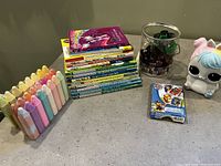 Children's books stacked, sidewalk chalk pieces in multiple colors, a Paw Patrol card set, plastic cutters in a container, and a small toy piggy bank on a countertop.