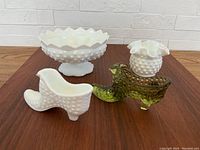 Front view of four Fenton hobnail glass pieces on wooden table against white brick backdrop, showing a white hobnail glass large vase bowl, white hobnail shoe vase, green hobnail shoe vase, and white hobnail candlestick holder.