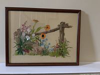 Framed embroidery picture showing a rustic wooden fence with colorful wildflowers on fabric canvas.